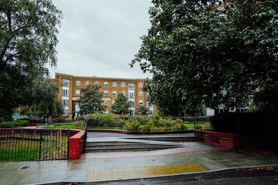 Photograph of an apartment building situated at the end of a short, paved pathway with a gentle incline, surrounded by a low metal fence with red brick columns. The pathway leads from a sidewalk that