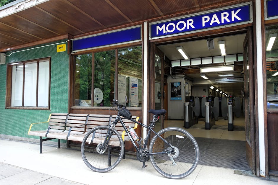 The image shows the exterior entrance of Moor Park station with a blue sign displaying the station name. In front of the station, there is a wooden bench with metal armrests and a black bicycle parked beside it, secured with a lock. To the left, the building's wall is covered in green tiles with a small yellow RVP sign near a large window that reveals some interior posters and information notices. The entrance to the station features automated ticket gates with waist-high barriers, and inside, fluorescent lights illuminate a long corridor. The environment appears clean and organized, suitable for home relocation or furniture transport services provided by Man with Van Raynes Park, with the station serving as a key route for efficient moving logistics and packing and moving operations.