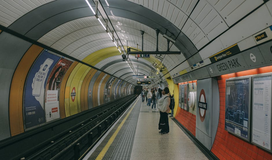 A fleet of modern train carriages parked on multiple railway tracks visible from an overhead perspective at Raynes Park station. Several trains are positioned on adjacent tracks, with two yellow and grey train units facing forward, one on the right side and another slightly behind on the central track. The train units are connected to overhead electric wires supported by tall metal poles and gantries. The surrounding environment features a mix of residential and commercial buildings, some under construction, in the background, with leafless trees indicating colder weather. The tracks are laid on gravel beds, with safety barriers and fencing along the platform edges on the right, helping to manage movement and safety during operations. The sky appears overcast, casting diffuse lighting across the scene. This image reflects railway infrastructure, crucial for home relocation logistics that involve strategic transportation planning, with [COMPANY_NAME] ensuring professional moving and furniture transport services aligned with such transit routes from Raynes Park station.