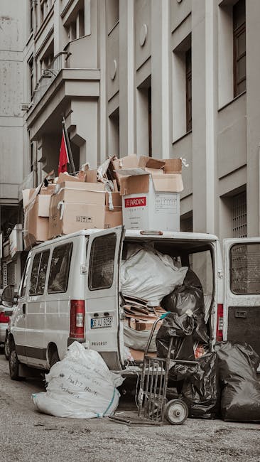 A white commercial van used for furniture transport and home relocation is parked on a city street adjacent to a multi-storey building with a grey façade and large windows. The van's rear doors are open, revealing an interior filled with large plastic bags, cardboard boxes, and wrapped furniture items prepared for moving. Several black and white garbage bags and flattened cardboard boxes are either inside or stacked at the back of the vehicle, indicating recent packing or packing materials used during packing and moving. The load on top of the van consists of open cardboard boxes with packing materials such as paper and foam, some of which appear to be in the process of being loaded or unloaded. A small wheeled trolley is positioned nearby, ready for transport of heavier items. The scene portrays the moving process, likely during a home relocation, with the focus on packing, loading, and furniture transport logistics, supported by the presence of packing materials and moving equipment consistent with professional removals by Man with Van Raynes Park.