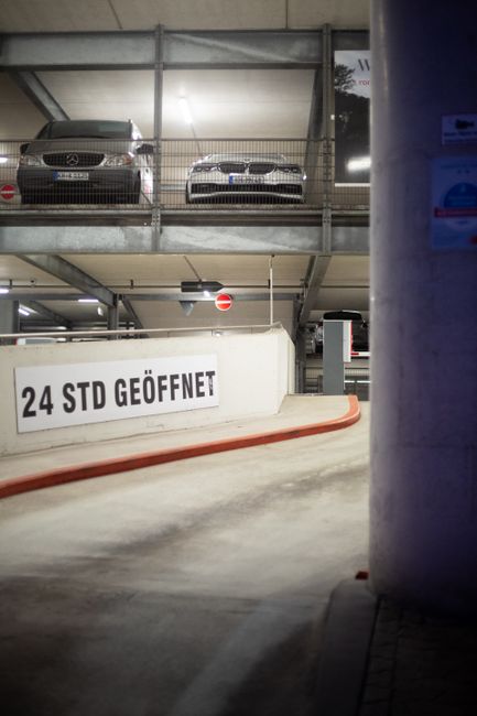 Inside a multi-storey car parking garage with a curved ramp leading to the upper level where two cars are parked; one silver Mercedes-Benz and one white BMW, both positioned facing forward. The parkin