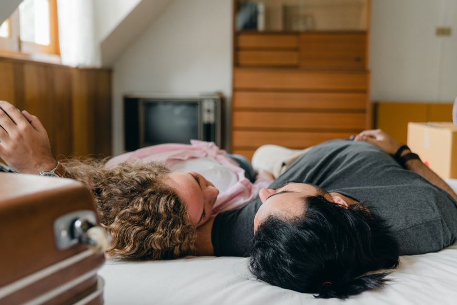 A woman and a man lying on a bed inside a room filled with cardboard boxes and packing materials. The woman is resting her head on her hand, appearing concerned or thoughtful, while the man, lying nex