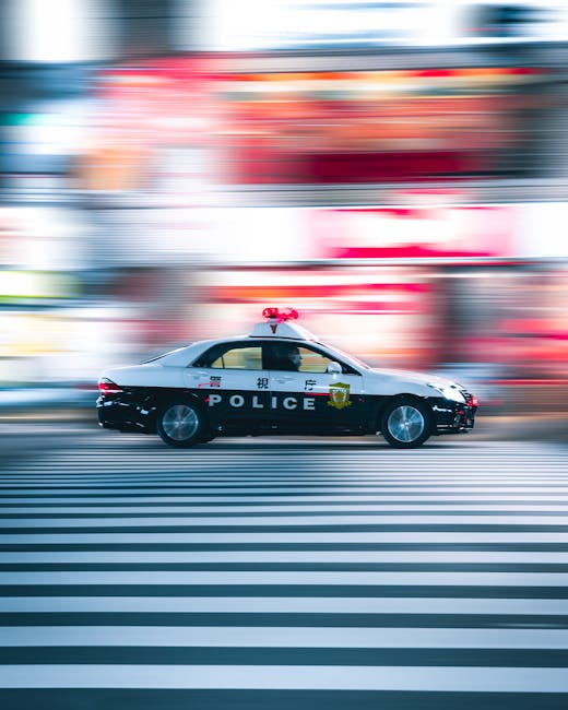 A black and white police car with red and blue lights on the roof, moving quickly along a city street with a motion blur effect. The vehicle features police insignia and the word 'POLICE' on the side. The background shows red and beige building facades with signage and storefronts, while the road has painted zebra crossing lines. The scene captures a fast-paced urban environment, likely during daytime, as part of a dynamic scene related to emergency response or city transportation. This image is relevant to house removals and relocation logistics, illustrating the importance of transportation and timely movement of services, as seen in the context of emergency or urgent moves managed by companies like Man with Van Raynes Park.