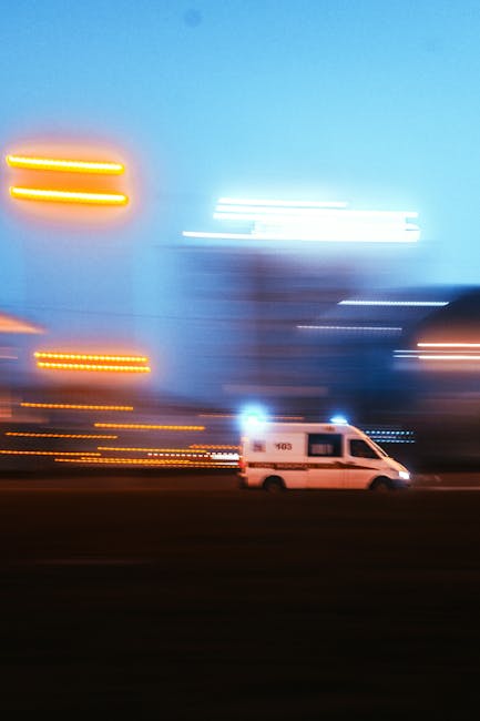 A white van from Man with Van Raynes Park is shown in motion on a city street during dusk or evening, with blurred streaks of orange, yellow, and white city lights creating a sense of rapid movement and dynamic environment. The van appears to be part of a home relocation or furniture transport service, with the surrounding urban setting indicating movement between locations. The image captures the loading or transport process characteristic of professional removals, emphasizing the speed and efficiency of emergency same-day moves in SW20. Streetlights and building lights in the background are blurred, highlighting the focus on the van in transit, which is positioned centrally on the road, moving from left to right, with the cityscape fading into a blue sky. This visual aligns with the logistical aspects of house removals, packing, and furniture transport facilitated by Man with Van Raynes Park.