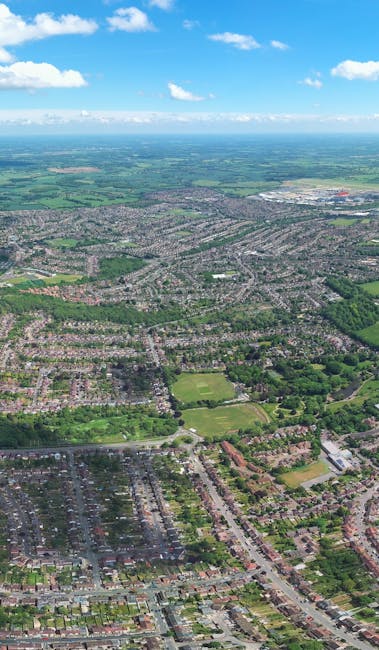 An aerial view of a suburban neighbourhood during daytime, showing rows of terraced and detached houses with rooftops primarily in shades of grey and brown. Streets are lined with trees and greenery, including parks and open fields scattered throughout the area. In the foreground, residential streets extend towards the horizon, with some areas featuring parked cars along the curbs. The landscape transitions from densely built housing to more open green spaces and farmland in the distance, under a bright blue sky with a few scattered clouds. The image illustrates the expansive scope of a home relocation or furniture transport operation, which Man with Van Raynes Park may facilitate, particularly in urban and suburban settings, supporting efficient packing, loading, and moving services. The scene emphasizes the importance of organized logistics in house removals within residential neighbourhoods.