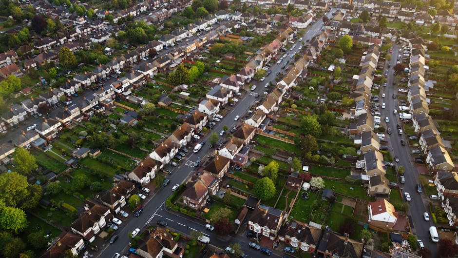 An aerial view of a residential neighbourhood showing multiple rows of detached and semi-detached houses with individual gardens. The houses are arranged along curved and straight streets, with cars parked along the curbs and within driveways. The gardens are lush with green lawns, trees, and bushes, some with fences or hedges separating properties. The roofs vary in color, including shades of brown and grey, and some houses have visible chimneys and small porches. The environment appears quiet and orderly, with well-maintained streets, and the image captures the daytime with natural lighting. This scene illustrates a typical UK suburb suitable for house removals and home relocation services provided by Man with Van Raynes Park, aiming at efficient furniture transport and packing logistics in a residential area.