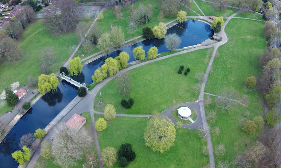 An aerial view of a park featuring a winding river with a small arched stone bridge crossing over it at the center. The park is surrounded by lush green grass, various mature trees, and pathways that radiate outward, some of which are empty while others are bordered by neatly clipped bushes. A small white pavilion is located on a circular grassy area in the lower part of the image, near the middle of the park. To the left of the river, there is a single-story brick building with a pitched roof, partially obscured by trees. The landscape is evenly lit with natural daylight, highlighting the contrasting colors of the foliage and water. This scene depicts a tranquil outdoor environment suitable for leisure walks and outdoor activities, typical of a public park in Raynes Park, and aligns with the context of house removals and moving logistics, with the open spaces potentially used for relocating items or as part of a moving process supported by Man with Van Raynes Park.