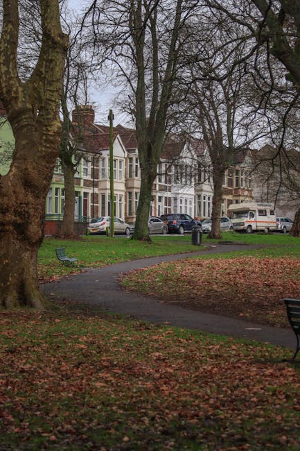 A paved pathway winds through a small, grassy park area with fallen leaves scattered across the ground. Several large, leafless trees with thick trunks are present, indicating a late autumn or early winter setting. In the background, residential houses with bay windows and pitched roofs are visible, along with parked cars on the street, including a white van which may be associated with [COMPANY_NAME] for local house removals. The scene is outdoors during daylight hours with overcast sky, and there is a blue park bench near the pathway, emphasizing the park environment. This setting reflects a typical urban or suburban area where furniture and belongings are often transported during home relocation, with the van potentially used for loading or unloading household items as part of a moving or packing process.