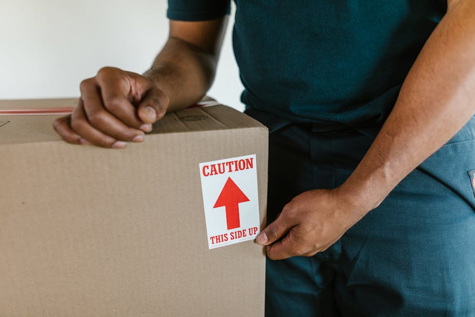A person dressed in a dark green T-shirt and dark trousers is holding a medium-sized cardboard box with a red and white warning sticker that reads 'Caution This Side Up' with an upward-pointing arrow. The individual is gripping the box with both hands, preparing to load or transport it within a home environment. The background shows an indoor setting with neutral walls, indicating a typical room during a house removal process. The box appears sealed with packing tape, and the action suggests careful handling as part of packing or furniture transport during a home relocation. This scene exemplifies the packing and handling aspects involved in professional removals, supported by the services of [COMPANY_NAME], which specialises in house removals and moving logistics.