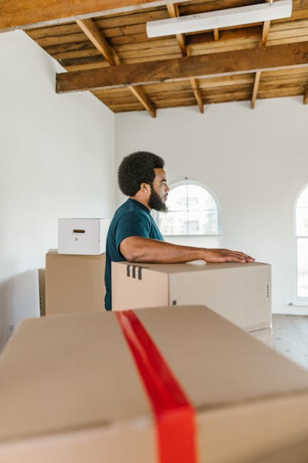 A person dressed in a dark green T-shirt and dark trousers is holding a medium-sized cardboard box with a red and white warning sticker that reads 'Caution This Side Up' with an upward-pointing arrow. The individual is gripping the box with both hands, preparing to load or transport it within a home environment. The background shows an indoor setting with neutral walls, indicating a typical room during a house removal process. The box appears sealed with packing tape, and the action suggests careful handling as part of packing or furniture transport during a home relocation. This scene exemplifies the packing and handling aspects involved in professional removals, supported by the services of [COMPANY_NAME], which specialises in house removals and moving logistics.
