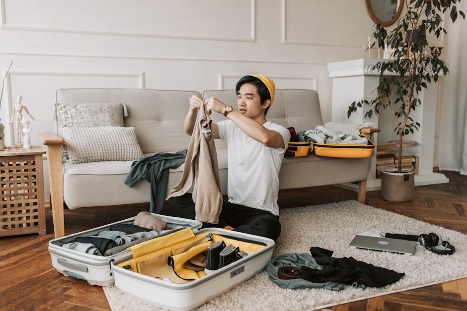 A young man with light skin and dark hair, wearing a white t-shirt, black trousers, and a yellow headband, is sitting on a cream-colored carpet inside a living room. He is in the process of packing clothes, holding a beige garment in both hands, preparing to place it into an open suitcase filled with assorted clothing and packing accessories. Surrounding him on the floor are additional clothing items, a pair of headphones, and a closed laptop. The suitcase nearby contains neatly rolled fabrics, a yellow strap, and various packing tools. Behind the young man, a light grey sofa with patterned cushions and a throw blanket is visible, along with a wooden side table holding decorative ornaments. To the right, an open yellow box filled with more folded clothes and packing materials rests on the sofa arm. The room features white paneled walls, a wooden floor, and a large potted plant near a fireplace mantel. This scene depicts home relocation and furniture transport preparations, aligning with packing and moving processes carried out by Man with Van Raynes Park.