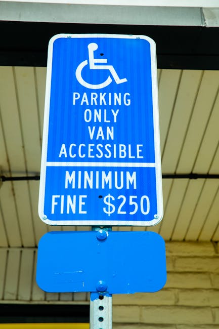 A blue rectangular parking sign mounted on a metal pole located beneath a ceiling or roof overhang. The sign features a white wheelchair symbol at the top, followed by the text 'PARKING ONLY VAN ACCESSIBLE' in white capital letters. Below that, there is a white horizontal line, and the words 'MINIMUM FINE $250' are printed at the bottom. The background of the image shows part of a building structure with beige and cream-colored walls and a ceiling with horizontal paneling. The sign indicates designated parking for vans accessible for individuals with mobility impairments, which is relevant to the context of home relocations and moving services provided by Man with Van Raynes Park, especially during parking suspensions and fines associated with SW20 moves.
