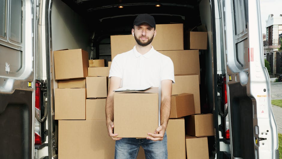 A man wearing a black cap and a white polo shirt is standing outside near the open rear doors of a white moving van. He is holding a cardboard box and smiling. Inside the van, there are several stacke