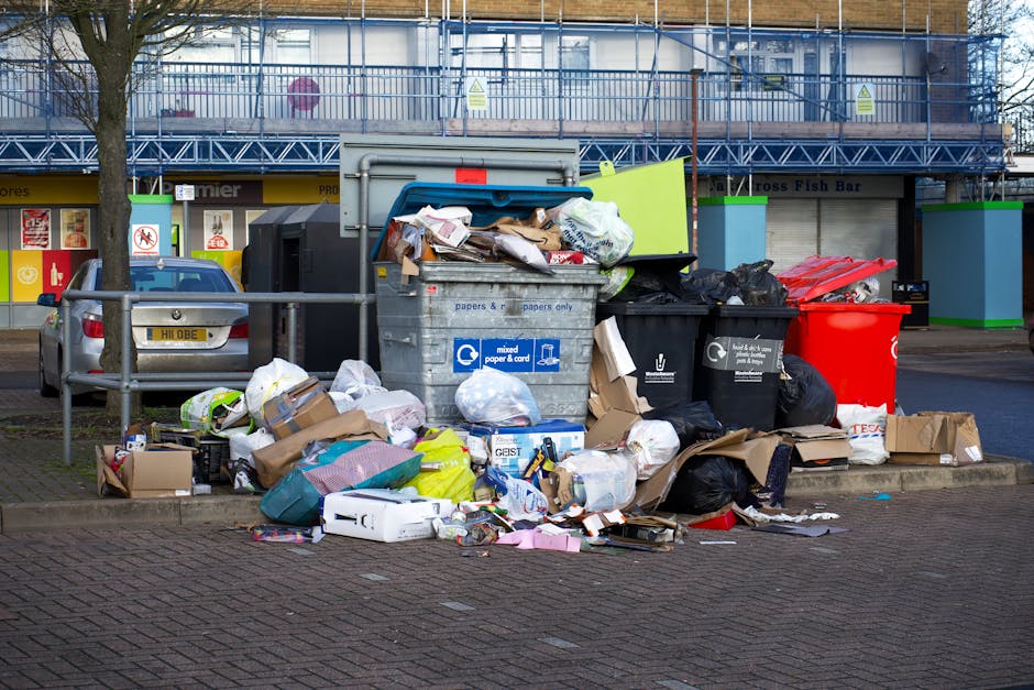 An outdoor scene of a public rubbish disposal area in Raynes Park featuring multiple overflowing waste bins and surrounding litter. The central focus is a large grey mixed paper and cardboard recycling bin filled to capacity, with brown cardboard boxes, plastic bags, and paper scattered on the pavement around its base. To the left is a black bin with a partially open lid, containing black waste bags, alongside a red waste container, both positioned on the sidewalk. To the right of the recycling bin, there is a black bin with a closed lid, also filled with waste, and additional black trash bags stacked nearby. Behind the bins, a metal railing separates the waste area from a street, where a parked silver car with a visible license plate is situated. In the background, a building with retail storefronts displaying signs, including a fish bar, and a blue scaffolding structure suggests ongoing construction or maintenance. The overall scene depicts improper waste disposal typical of house removal or moving-related debris, supporting the topic of rubbish disposal regulations in Raynes Park. The image highlights the importance of correct waste management for home relocations, with visible materials such as plastic, paper, cardboard, and fabric waste scattered around the bins, emphasizing the need for appropriate clearance services provided by Man with Van Raynes Park.