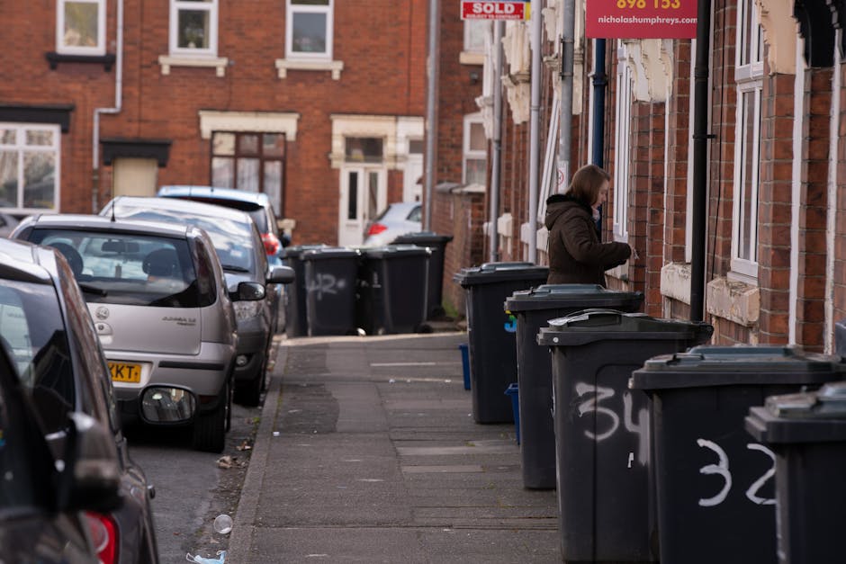 An outdoor scene of a public rubbish disposal area in Raynes Park featuring multiple overflowing waste bins and surrounding litter. The central focus is a large grey mixed paper and cardboard recycling bin filled to capacity, with brown cardboard boxes, plastic bags, and paper scattered on the pavement around its base. To the left is a black bin with a partially open lid, containing black waste bags, alongside a red waste container, both positioned on the sidewalk. To the right of the recycling bin, there is a black bin with a closed lid, also filled with waste, and additional black trash bags stacked nearby. Behind the bins, a metal railing separates the waste area from a street, where a parked silver car with a visible license plate is situated. In the background, a building with retail storefronts displaying signs, including a fish bar, and a blue scaffolding structure suggests ongoing construction or maintenance. The overall scene depicts improper waste disposal typical of house removal or moving-related debris, supporting the topic of rubbish disposal regulations in Raynes Park. The image highlights the importance of correct waste management for home relocations, with visible materials such as plastic, paper, cardboard, and fabric waste scattered around the bins, emphasizing the need for appropriate clearance services provided by Man with Van Raynes Park.