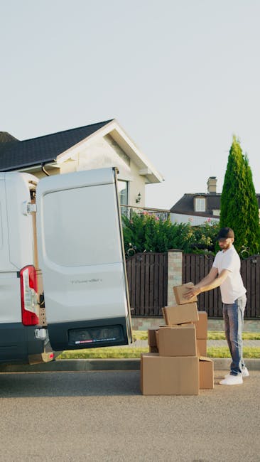 A man wearing a white t-shirt, jeans, white sneakers, and a black cap is engaged in a home relocation task outside a residential property. He is carrying three medium-sized cardboard boxes stacked one