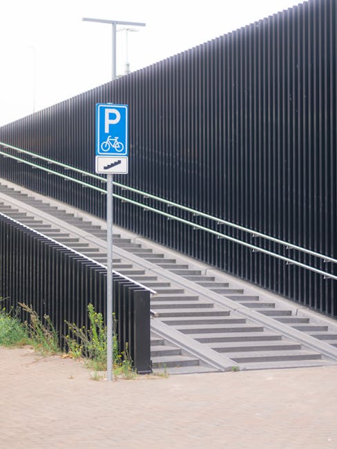 The image shows a set of outdoor metal stairs with multiple steps leading upward, adjacent to a tall, dark vertical fence made of slim metal slats. The stairs are crossed by a handrail on each side, painted in a light color, and appear to be designed for pedestrian and bicycle access. To the left of the stairs, there is a blue parking sign with a white bicycle icon, indicating bicycle parking or access, mounted on a metal pole. The ground in front of the stairs and fence is paved with light-colored bricks or concrete, with some small green plants growing near the base of the fence. The background includes a cloudy sky with partial view of a television or communication antenna. This outdoor scene depicts an urban or suburban environment, possibly part of a housing or commercial complex, relevant to logistics or moving services such as those offered by Man with Van Raynes Park for staircase and narrow-access house moves.