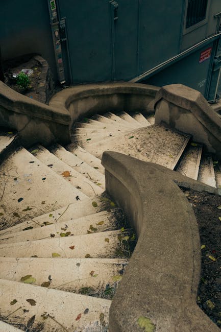 A curved outdoor stone staircase leading down from a building, with worn, slightly stained steps covered in scattered fallen leaves. The staircase features a smooth, rounded concrete handrail on either side, guiding the descent. At the top of the stairs, part of a blue-grey metal door or gate is visible, indicating the entrance to a property, with some greenery nearby. The setting appears to be an urban or residential area, suggesting this staircase may be used during home relocation or moving processes, with the environment illuminated by natural daylight. This image, associated with Man with Van Raynes Park, reflects the challenging access typical of staircase and narrow-access moves around Cannon Hill, RAYNES PARK, and highlights the importance of careful handling during furniture transport and home removals, especially in confined or complex stairwell environments.