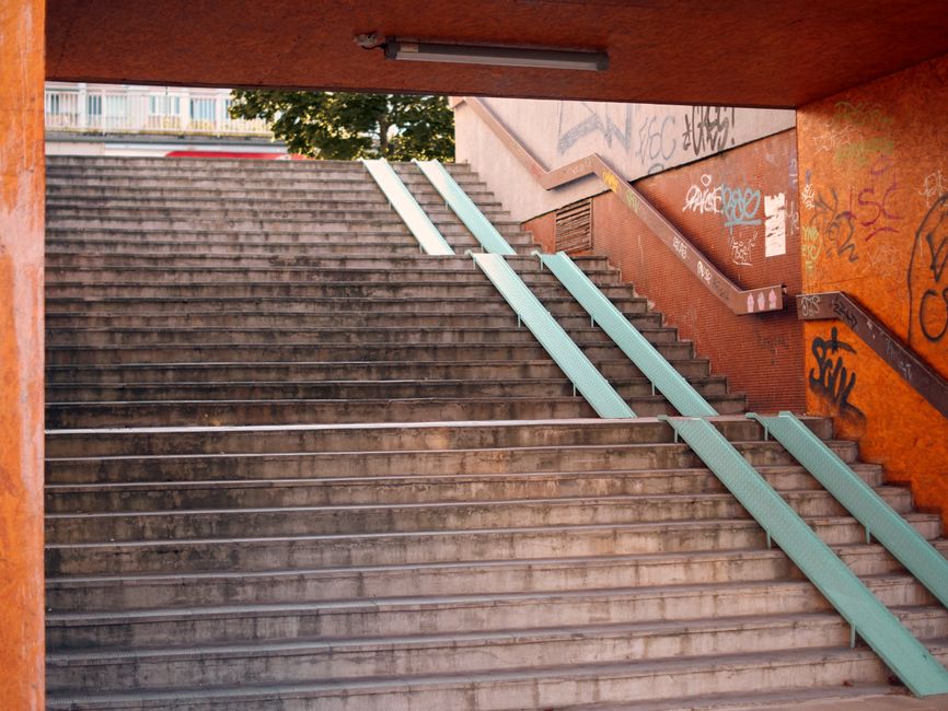 The image shows a set of outdoor metal stairs with multiple steps leading upward, adjacent to a tall, dark vertical fence made of slim metal slats. The stairs are crossed by a handrail on each side, painted in a light color, and appear to be designed for pedestrian and bicycle access. To the left of the stairs, there is a blue parking sign with a white bicycle icon, indicating bicycle parking or access, mounted on a metal pole. The ground in front of the stairs and fence is paved with light-colored bricks or concrete, with some small green plants growing near the base of the fence. The background includes a cloudy sky with partial view of a television or communication antenna. This outdoor scene depicts an urban or suburban environment, possibly part of a housing or commercial complex, relevant to logistics or moving services such as those offered by Man with Van Raynes Park for staircase and narrow-access house moves.